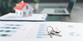 A set of house keys laying on top of paperwork with a house replica behind them.